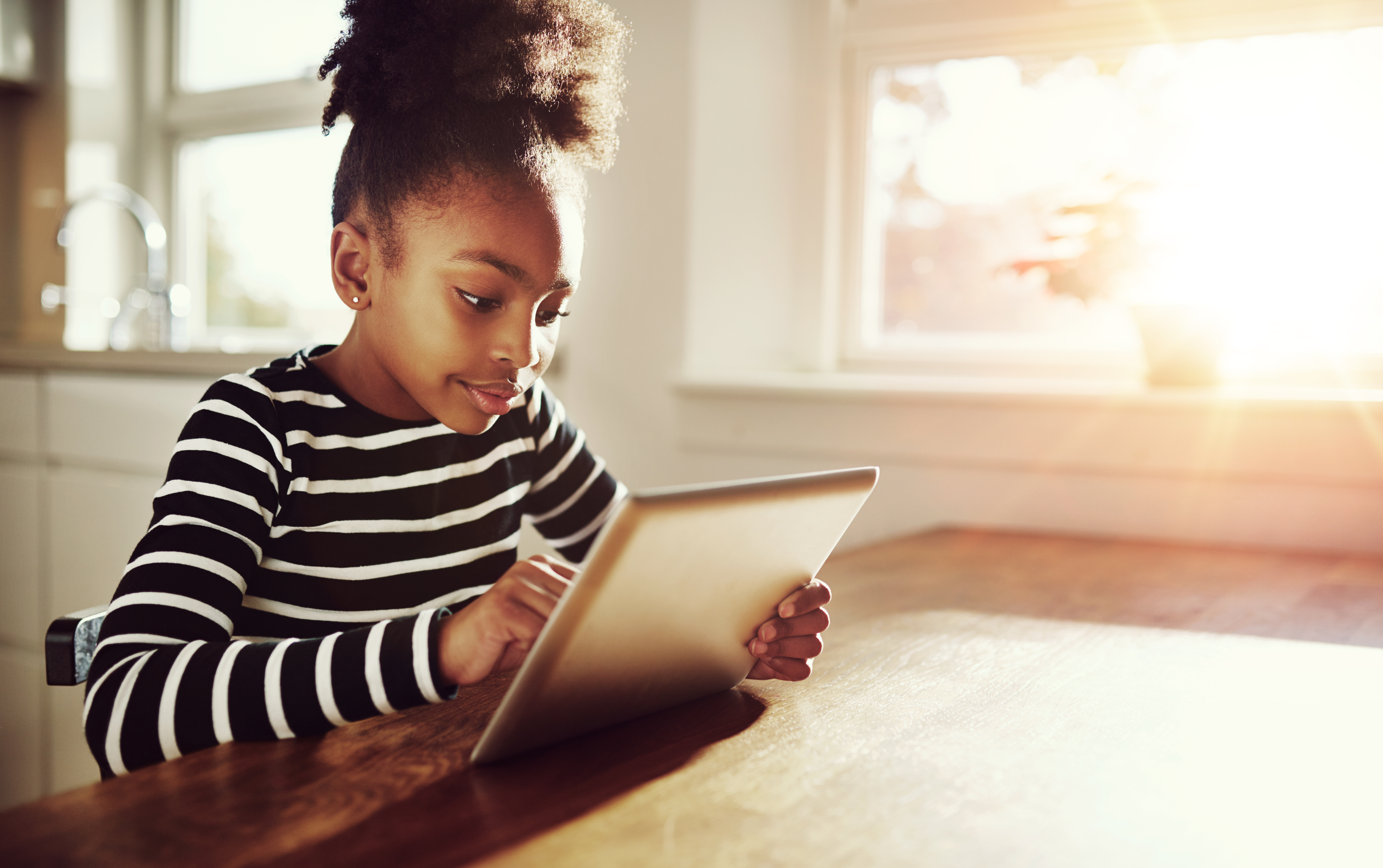 Young black girl with a fun afro hairstyle sitting at a table at home browsing the internet on a tablet computer with bright sun flare through the window alongside her